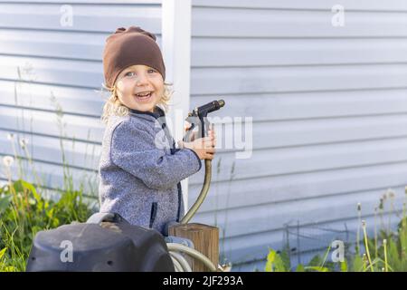 Nahaufnahme eines fröhlichen kleinen Jungen unter fünf Jahren, der im Hinterhof steht und die Spritzpistole eines Gartenschlauchs mit Kopierraum nach rechts hält. Stockfoto