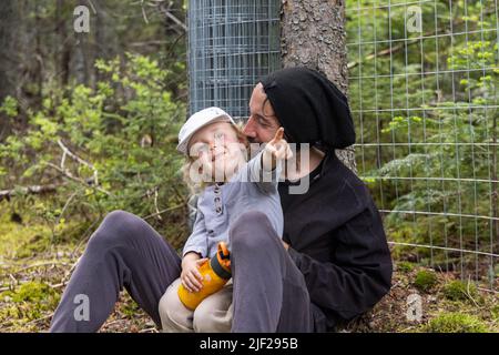 Glücklicher Vater und junger Sohn ruhen sich in der Natur aus. Der zweijährige Junge sitzt auf dem Schoß des Vatis und zeigt lächelnd mit dem Finger, mit verschwommenem Wald im Hintergrund. Stockfoto