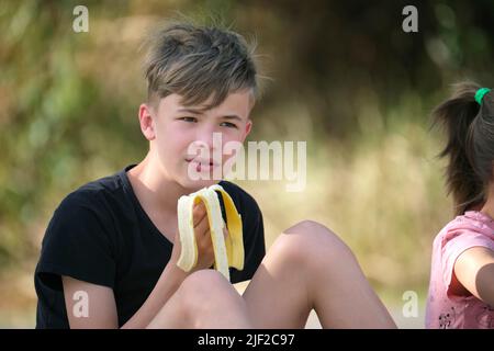 Junger hübscher Teenager Junge, der am Sommertag leckere reife Bananen im Freien isst. Stockfoto