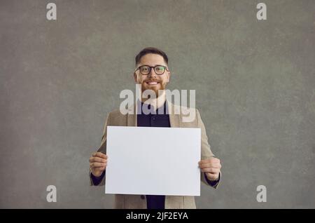 Junger, attraktiver, glücklicher Mann, der ein weißes, weißes Blatt in der Hand hält, während er auf einem grauen Hintergrund steht. Stockfoto