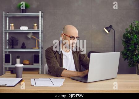 Ernsthafter Mann, der im Heimbüro am Schreibtisch sitzt und am Laptop tippt Stockfoto