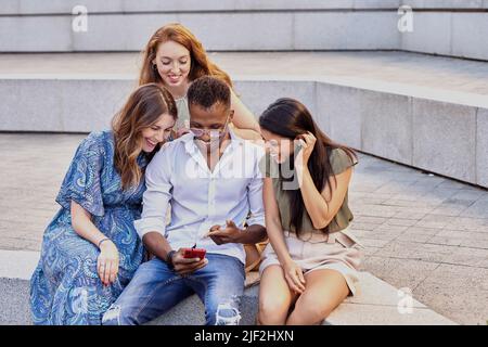 Gruppe von Freunden, die ein Mobiltelefon zusammen benutzen, während sie im Freien sitzen. Technologie, Social Media und Freundschaftskonzept. Stockfoto