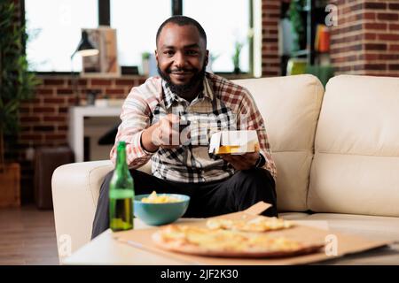Lächelnde Person wechselt mit der fernbedienung die Sender und isst Nudeln vom Essen zum Mitnehmen auf der Couch. Spaß mit Film oder Film, Essstäbchen zum Mitnehmen verwenden. Stockfoto