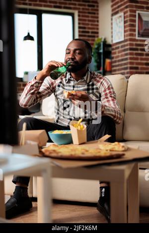 Glückliche Person trinkt eine Flasche Bier und isst Essen zum Mitnehmen von Fast-Food-Lieferung. Genießen Sie Essen zum Mitnehmen und schauen Sie sich Filme im Fernsehprogramm zu Hause an, essen Sie zu Abend und trinken Sie. Stockfoto