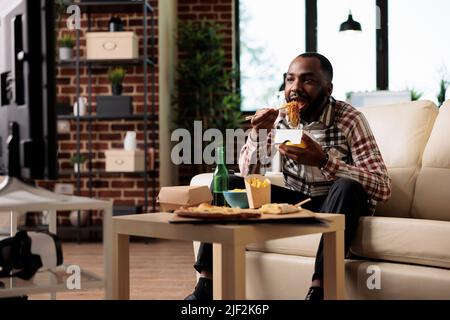 Zwanglose Person, die Nudeln aus der Lieferbox genießt und sich einen Film im Fernsehprogramm ansieht. Essen zum Mitnehmen mit Essstäbchen zur Unterhaltung, mit einer Flasche Bier. Stockfoto