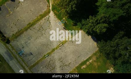 Ein altes Schulstadion in einem Stadtblock. Gesprungener Asphalt und Sportgeräte. Mehrstöckige Gebäude und eine große Grünfläche. Luftaufnahmen. Stockfoto