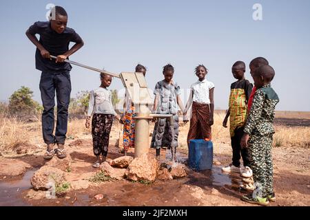 Mehrere junge afrikanische Dorfkinder gruppierten sich um eine manuelle Pumpe, die Kanister mit sauberem und frischem Trinkwasser füllt Stockfoto