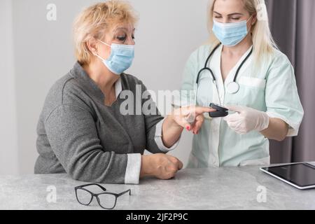 Hausgesundheitspflege. Frau Krankenschwester in einer medizinischen Maske helfen mittleren Alters Frau während der Krankheit oder Druck, weibliche Ärztin misst den Puls des Patienten Stockfoto
