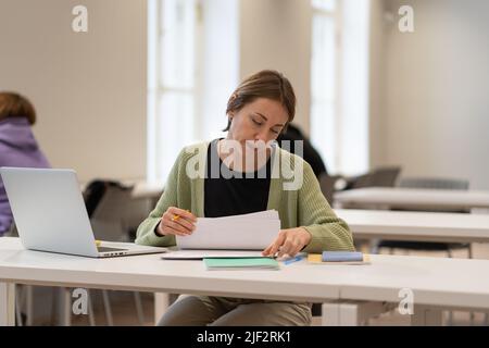 Fleißige reife Studentin, die Architekturzeichnungen liest, während sie in der öffentlichen Bibliothek studiert Stockfoto