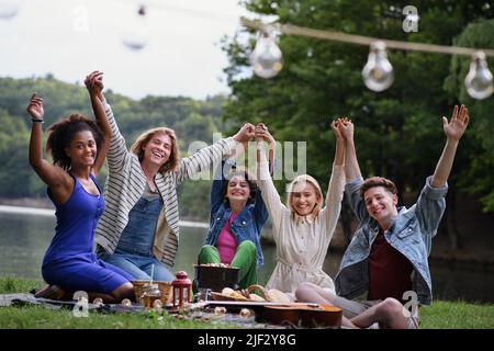 Eine Gruppe junger Freunde, die Spaß beim Picknick in der Nähe eines Sees haben, auf einer Decke sitzen und die Arme heben. Stockfoto