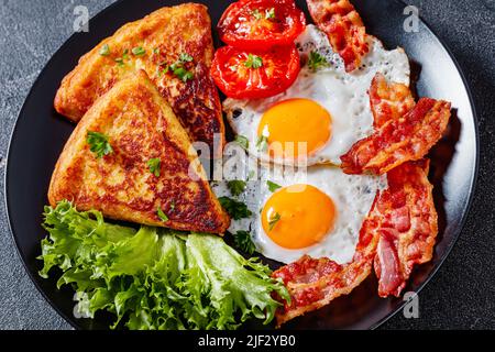 irisches Frühstück mit Spiegeleiern, gebratenen Speckscheiben, gebratenen Kartoffelkuchen, gegrillten Tomaten und frischem Salat auf schwarzem Teller auf Betontisch, Nahaufnahme Stockfoto