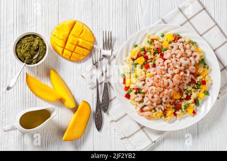 Gekochte kleine rosafarbene Garnelen auf Hirse-Pilaf mit grünen Erbsen, Mango, roten Paprika auf dem Teller auf weißem Tisch mit Pesto-Sauce und frischer Mango, hori Stockfoto