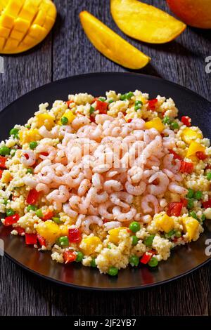 Gekochte kleine rosafarbene Garnelen auf Hirse-Pilaf mit Gemüse, Mangostückchen und Gewürzen auf schwarzem Teller auf dunklem Tisch, vertikale Ansicht von oben Stockfoto