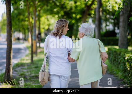 Rückansicht des Betreuers mit einer älteren Frau auf einem Spaziergang im Park mit Einkaufstasche. Stockfoto