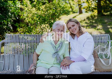 Pfleger mit einer älteren Frau, die im Sommer auf der Bank im Park sitzt und die Kamera anschaut. Stockfoto