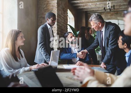 Zwei Geschäftsleute machen einen Deal durch Händeschütteln sitzen an einem Tisch zusammen mit anderen multirassischen Mitarbeitern - Business-Lifestyle und Vereinbarungen Konzept Stockfoto
