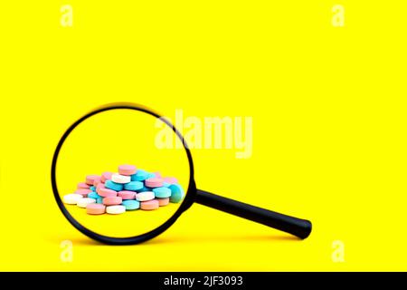 Ein Blick durch eine Lupe auf einen Haufen Pillen auf gelbem Hintergrund. Suchen Sie die Inhaltsstoffe von Medikamenten, Vitaminen, Antibiotika. Identifizieren Sie Fälschungen Stockfoto