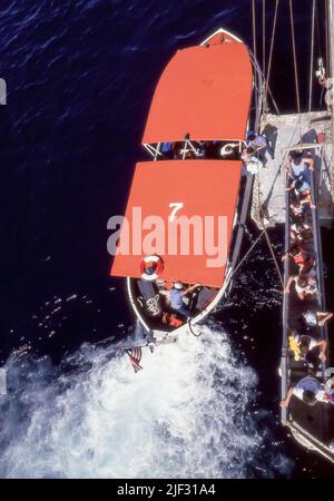 Gestartet von einem Ponton und einer über die Seite des Schiffes abgesenkten Gangway, bringt ein Tender (Rettungsboot) Passagiere nach einer Landreise an einem der Häfen, die das Kreuzschiff TSS Fairstar besucht, zurück zum Schiff. Diese Aufnahme wurde 1984 vom Promenadendeck aufgenommen und ja, das sind Rauch und Dämpfe vom Dieselmotor im Tender. Ursprünglich ein britisches Truppenschiff Oxfordshire wurde sie zum Liner Fairstar (Sitmar) umgebaut und reiste zwischen England und Australien bis 1974, als sie bis zu ihrer letzten Reise im Jahr 1997 Vollzeit-Südpazifik-Kreuzfahrten begann. Stockfoto