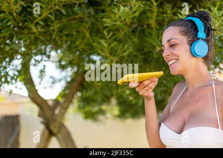 Lächelnde, hübsche junge Frau mit weißem Bikini und blauen kabellosen Kopfhörern sendet mit ihrem Mobiltelefon einen Ton, während sie sich am Pool sonnt Stockfoto