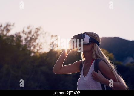 Frau, die bei Sonnenaufgang mit einer VR-Brille unterwegs ist, um die Metaverse zu erkunden. Blonde Frau auf Urlaub Wandern in einem Wald mit VR-Brille zu erleben erweitert Stockfoto