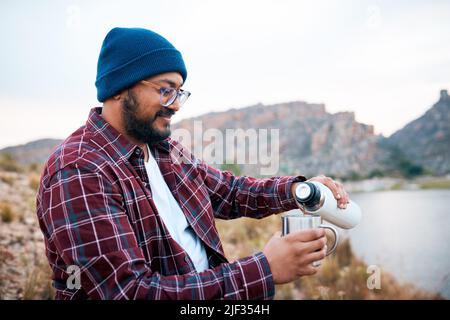 Ein erwachsener Mann gießt Kaffee auf einer Wanderung in den Bergen mit Seeblick Stockfoto