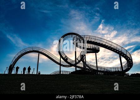 Fotografen mit Stativen als Silhouetten an der begehbaren Achterbahnskulptur Tiger and Turtle vor einem dunkelblauen Himmel mit weißen Wolken, Kunst ins Stockfoto