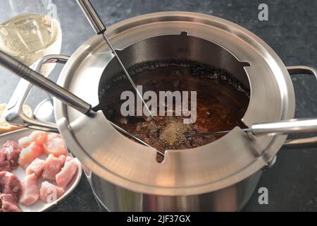 Blick in einen Fondue-Topf mit kochendem heißem Öl, langen Gabeln, Fleisch- und Weinstücken, festliches Abendessen für Partys mit Freunden und Familie, ausgewählter Fokus, Stockfoto