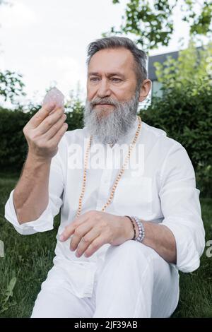 Leitender Meister-Guru in weißem Hemd und Perlen, der im Freien Selenitkristall ansieht Stockfoto