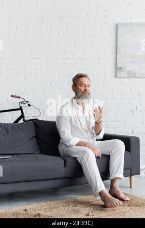 Älterer barfuß-Guru-Mann mit Mobiltelefon, der zu Hause auf der Couch sitzt Stockfoto