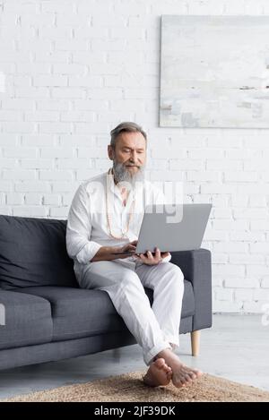 Bärtiger Meister-Guru in weißen Kleidern, der zu Hause mit einem Laptop auf dem Sofa sitzt Stockfoto
