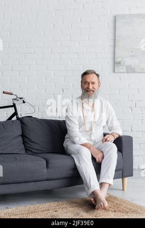Senior Master Guru in weißen Kleidern sitzt zu Hause auf dem Sofa und lächelt der Kamera zu Stockfoto