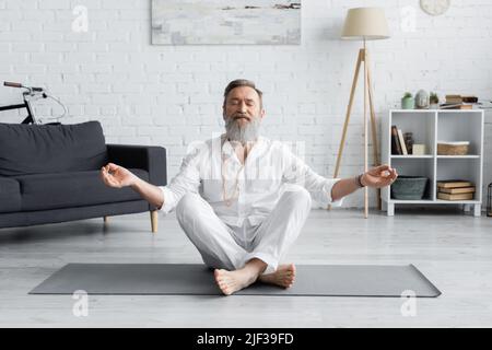 Bärtiger Guru-Trainer meditiert in leichter Pose mit geschlossenen Augen im Wohnzimmer Stockfoto