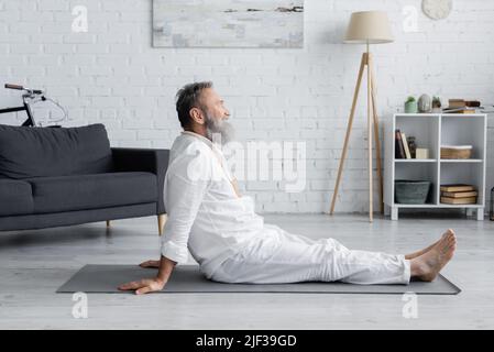 Seitenansicht des bärtigen Gurus-Mannes, der auf einer Yogamatte sitzt, während er zu Hause meditiert Stockfoto