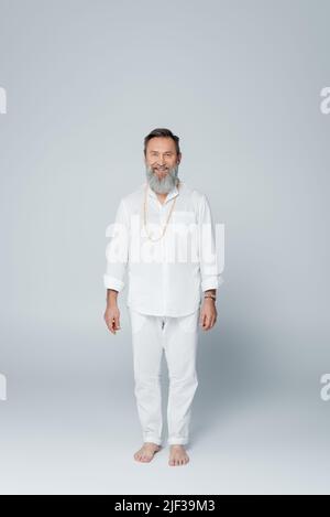In voller Länge lächelnder barfuß-Guru-Mann in weißen Kleidern, der auf grau steht Stockfoto