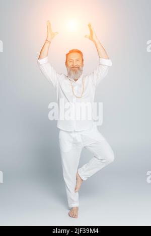 Die ganze Länge des Yoga-Gurus meditiert in Baumhaltung in der Nähe von leuchtendem Chakra auf grau Stockfoto