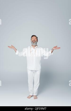 Die ganze Länge des lächelnden Yoga-Gurus meditiert in Baumhaltung mit offenen Armen auf grau Stockfoto
