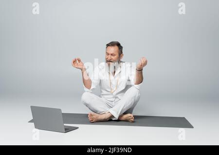 Grauhaariger Yogameister, der in leichter Pose sitzt und das Kinn-Mudra neben dem Laptop auf grau zeigt Stockfoto