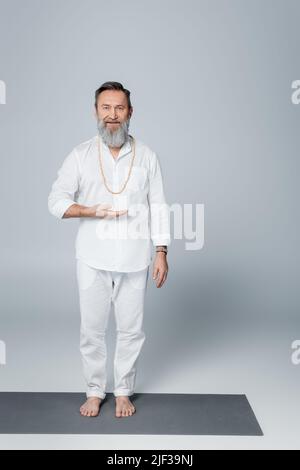 Leitender spiritueller barfuß-Guru zeigt mit der Hand und schaut auf die Kamera auf Grau Stockfoto