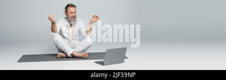 Senior Yoga Coach zeigt Kinnmudra in der Nähe des Laptops, während er in einer leichten Pose auf einem grauen Banner sitzt Stockfoto