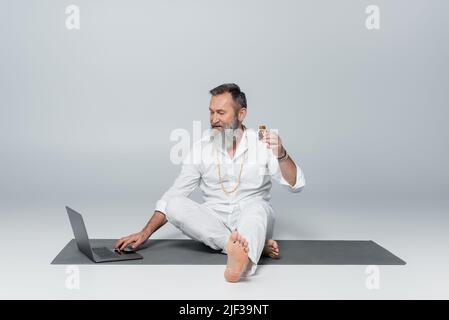 Barfuß heilender Guru sitzt neben dem Laptop und hält eine Flasche duftenden Öls auf grau Stockfoto