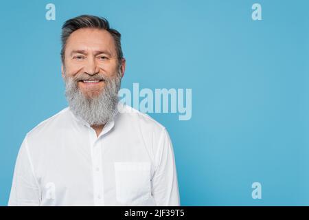 Glücklicher bärtiger Mann in weißem Hemd, der isoliert auf Blau auf die Kamera schaut Stockfoto