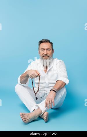 Bärtiger Barfußmann, der mit gekreuzten Beinen sitzt und mala-Perlen auf blauem Hintergrund hält Stockfoto