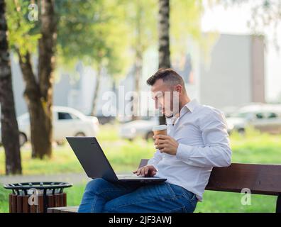 Ein ernsthafter Geschäftsmann arbeitet im Park mit einem Laptop und trinkt Kaffee. Junger Mann auf dem Hintergrund der vorbeifahrenden Autos, heißen sonnigen Sommertag. Warmes, weiches Licht, Nahaufnahme. Stockfoto