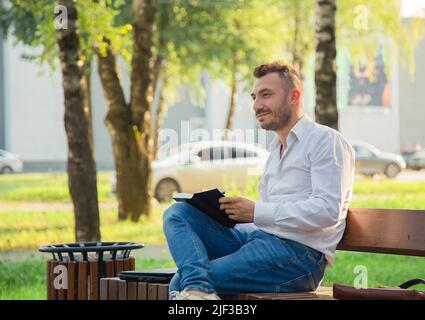 Ein glücklicher Mann sitzt auf einer Bank, macht Pläne, schreibt in ein Notizbuch. Ein junger Mann auf einem Hintergrund von grünen Bäumen, ein heißer sonniger Sommertag. Warmes, weiches Licht, Nahaufnahme. Stockfoto