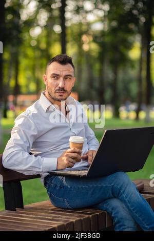 Serious Mann arbeitet im Park mit einem Laptop, trinkt Kaffee. Ein junger Mann auf einem Hintergrund von grünen Bäumen, ein heißer sonniger Sommertag. Warmes, weiches Licht, Nahaufnahme. Stockfoto