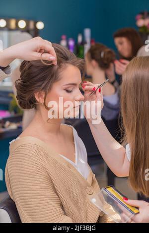 Die Hände der Frauen malen die Augen des Mädchens und lockieren ihr Haar im Salon. Make-up-Künstler und Friseur schaffen ein Bild einer jungen Frau. Geschäftskonzept - Schönheitssalon, Gesichtshaut- und Haarpflege. Stockfoto