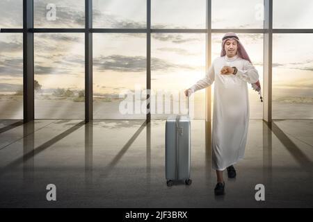 Arabischer Mann mit Keffiyeh und Blick auf die Uhr an seinem Handgelenk mit einem Koffer zum Reisen Stockfoto