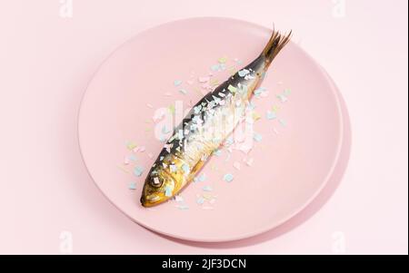 Fisch bedeckt Mikroplastik auf einem rosa Teller. Auswirkungen von Mikroplastik auf die Lebensmittelkette. Die Idee der Mikroplastik Verschmutzung. Konzept des Umweltdamms Stockfoto