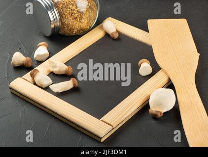 Verstreute Eringa-Pilze mit Menürahmen, Gewürz und Spatel auf schwarzem Hintergrund Stockfoto