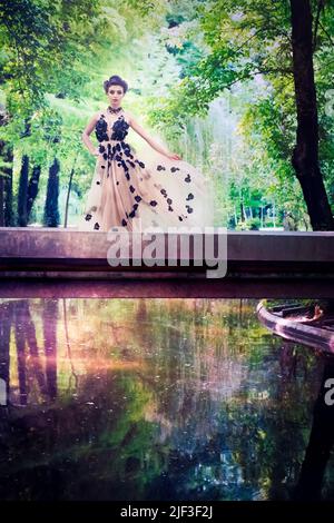 Schönes weibliches Modell mit langem Tüllblumenkleid und großen Haaren, die auf einer Brücke über buntes und helles Wasser in einem Park stehen. Konzept der bildenden Kunst Stockfoto